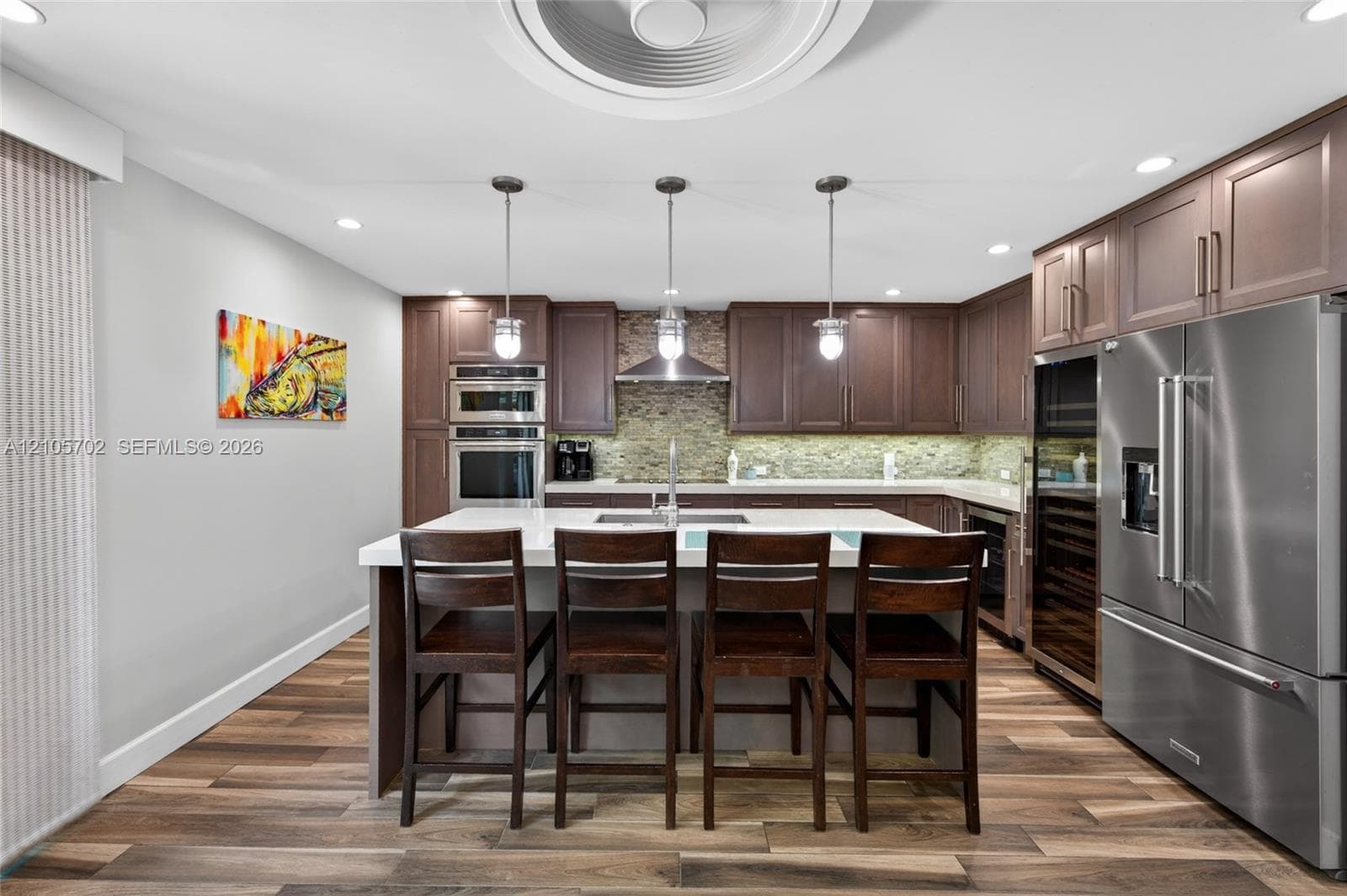 Kitchen featuring wood-finish cabinetry, stone-tiled backsplash, quartz countertops, a central island with integrated sink, and stainless steel appliances including a built-in wall oven