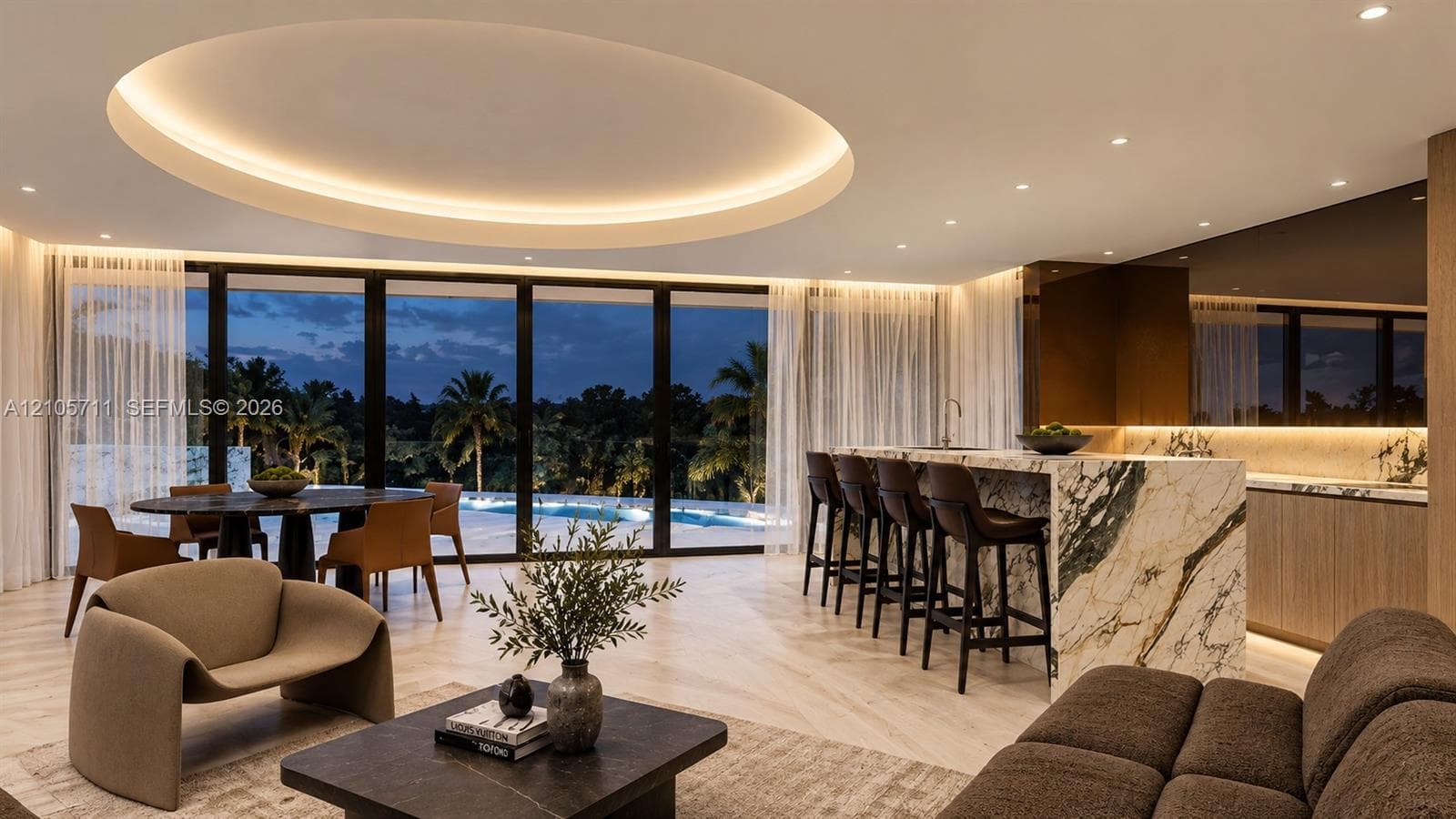 Expansive interior featuring a recessed circular tray ceiling with integrated perimeter lighting, floor-to-ceiling glass wall, light-toned wood-finish flooring, and a large kitchen island with a stone-surfaced waterfall edge