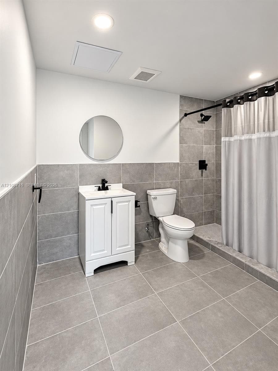 Bathroom featuring grey large-format tile flooring and wall accents, white vanity with a rectangular sink, circular wall-mounted mirror, and matte black fixtures