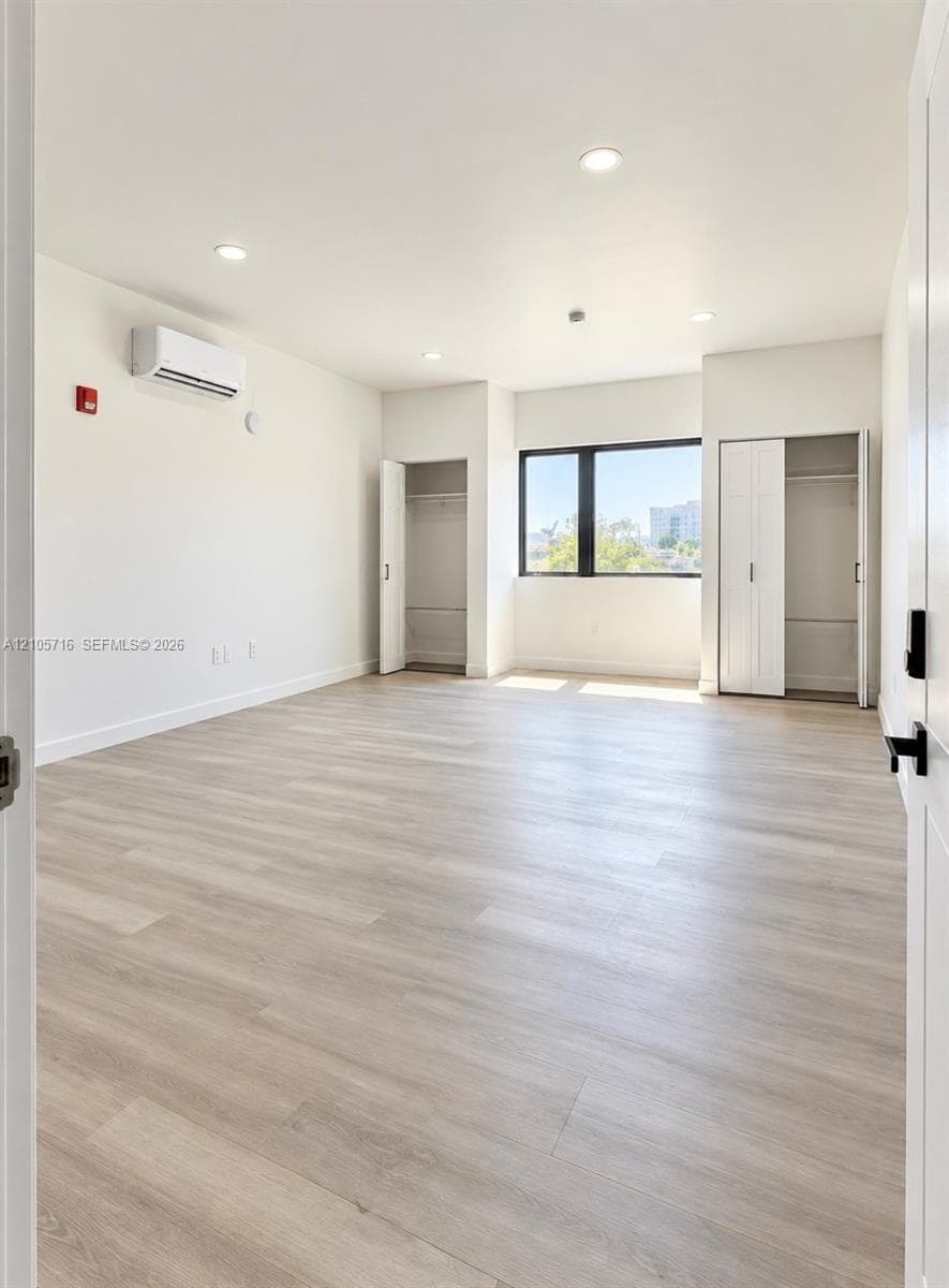 Neutral-toned room featuring wood-finish flooring, recessed lighting, dual closets with biflaminar doors, and a wall-mounted climate control unit