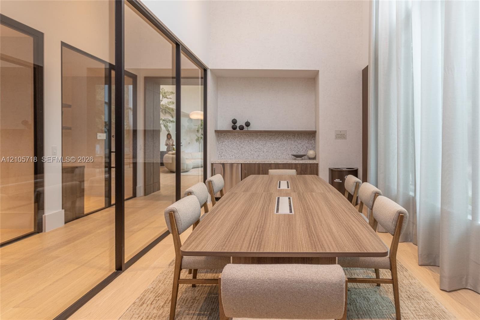 High-ceilinged room featuring expansive glass wall partitions with black metal frames, integrated built-in cabinetry with stone-surface counters, and light-toned wood-finish flooring