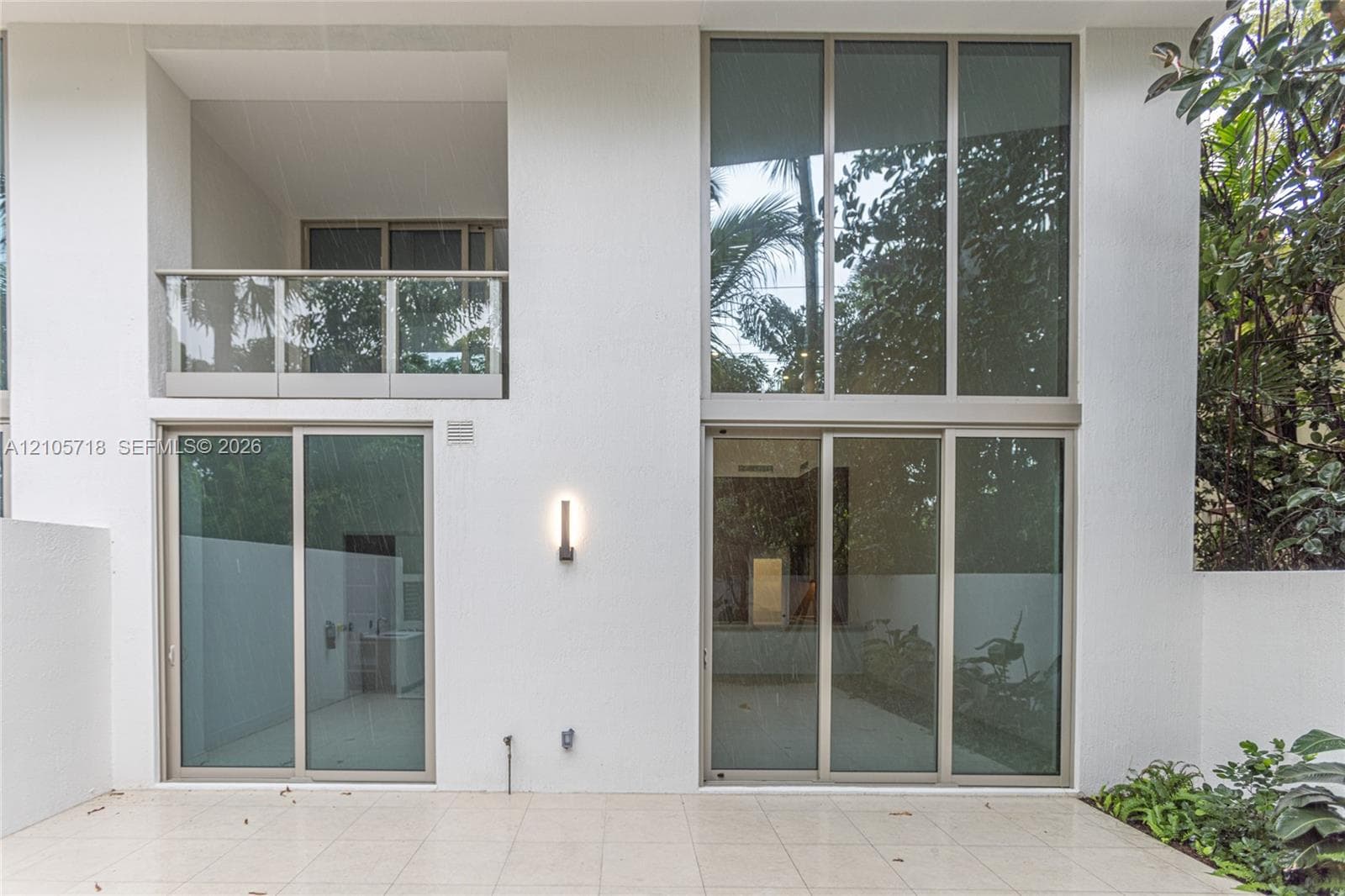 Exterior elevation featuring expansive two-story floor-to-ceiling windows, a covered balcony with a glass railing, large-format tile patio flooring, and wall-mounted minimalist lighting