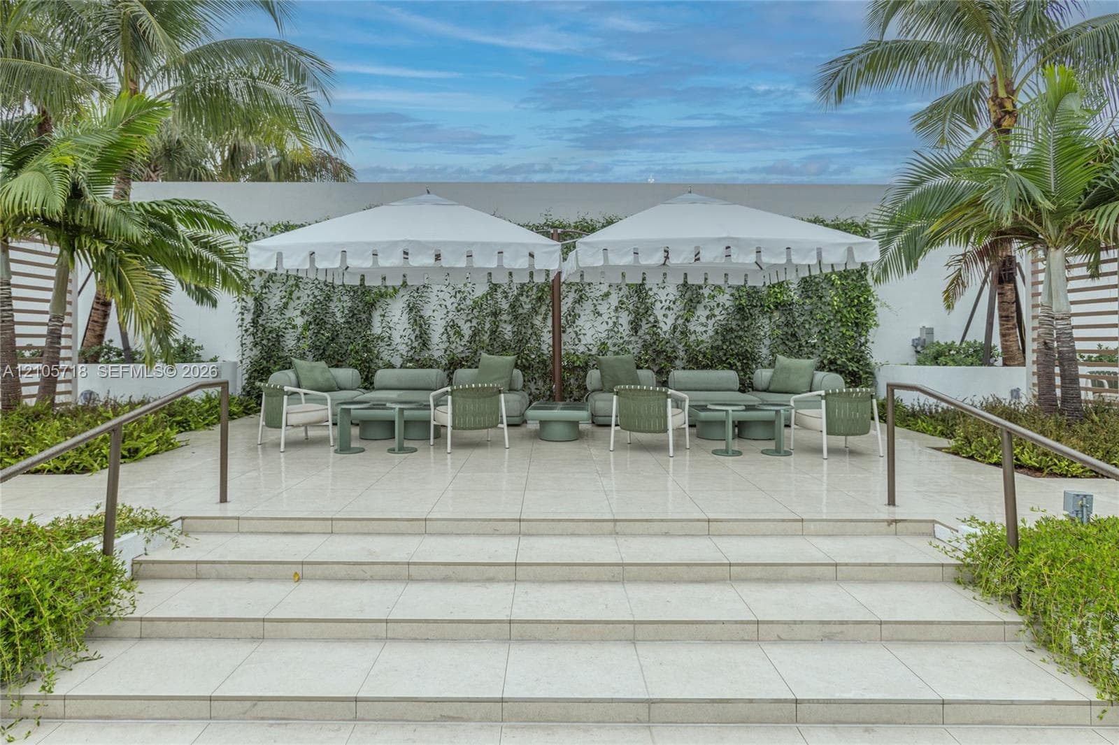 Outdoor terrace featuring oversized light-colored tiling, a tiered staircase with metal handrails, a climbing vine wall, and perimeter tropical landscaping