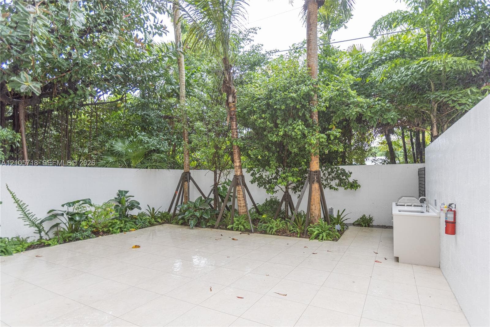 Outdoor courtyard featuring large-format tile flooring, built-in masonry counter with sink, solid perimeter wall, and established landscaping with mature palm trees