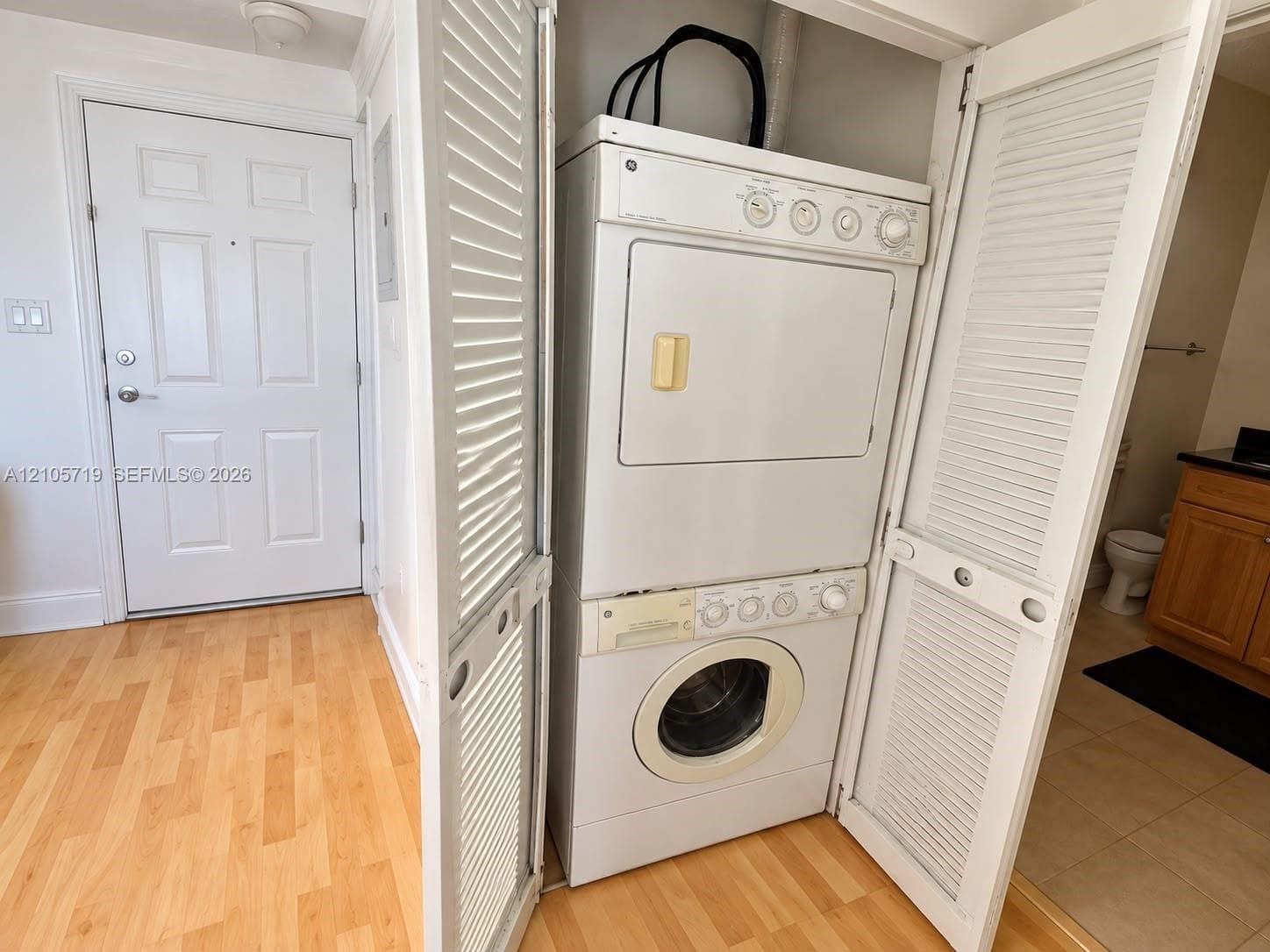 Stacked laundry unit housed in a louvered-door closet featuring wood-finish flooring and a multi-panel interior door
