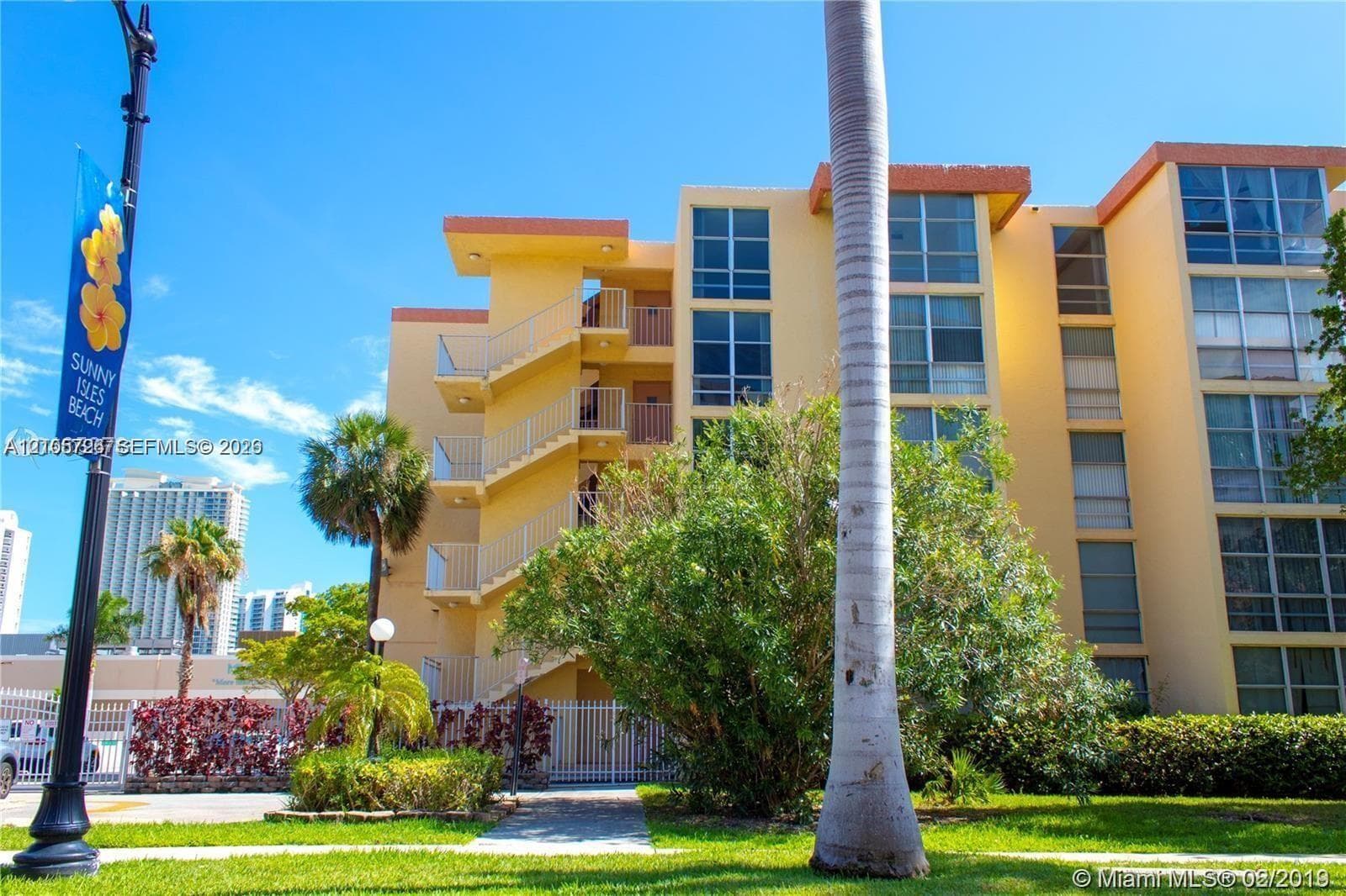 Multi-story residential building exterior featuring a stucco facade, expansive glass windows, external staircases with metal railings, and landscaped grounds with mature palm trees