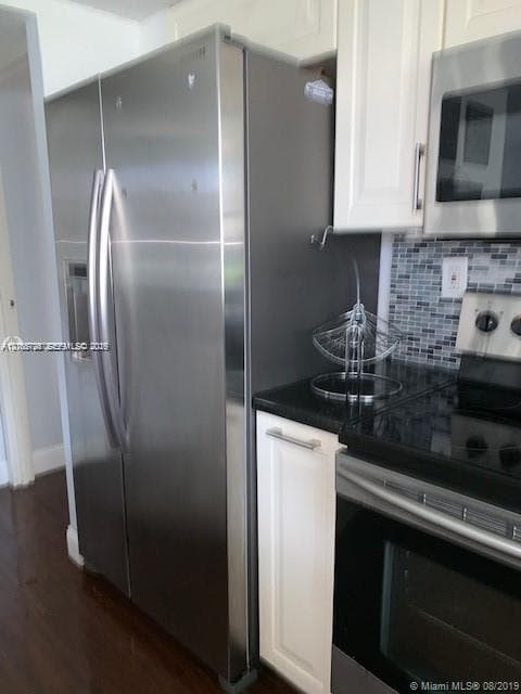 Stainless steel side-by-side refrigerator, white shaker-style cabinetry, dark solid-surface countertop, glass mosaic tile backsplash, and stainless steel built-in microwave