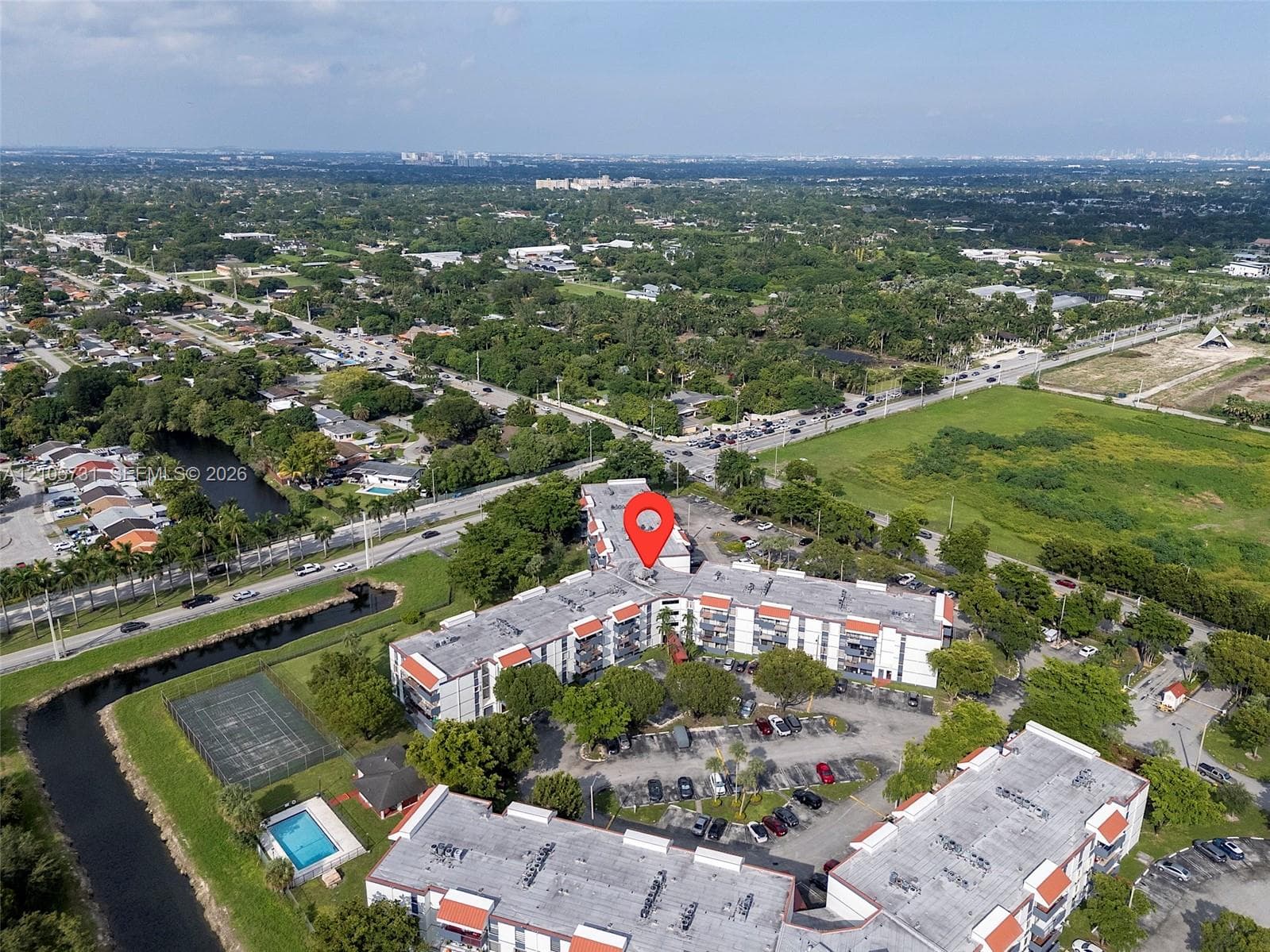 Aerial perspective of a multi-story residential building featuring integrated surface parking, an outdoor swimming pool, and a hardscaped tennis court