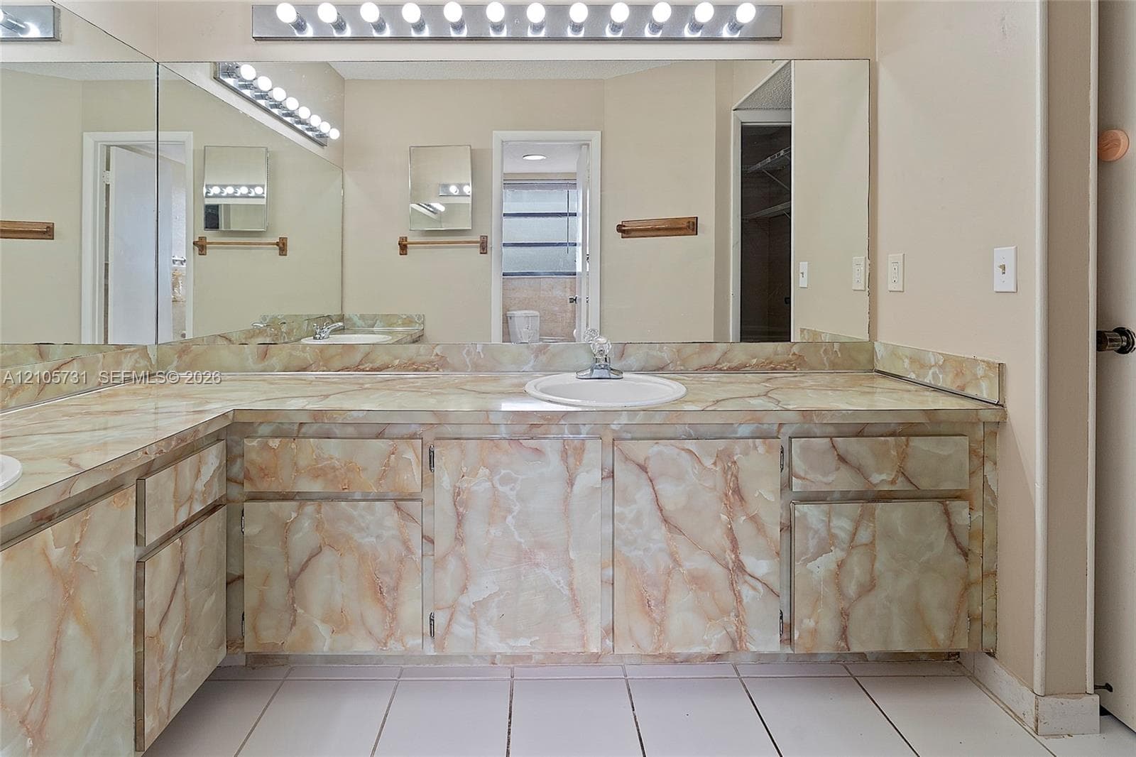 Double vanity featuring natural stone-patterned cabinetry and matching stone countertop with drop-in sinks, wall-to-wall mirror, and overhead multi-bulb lighting
