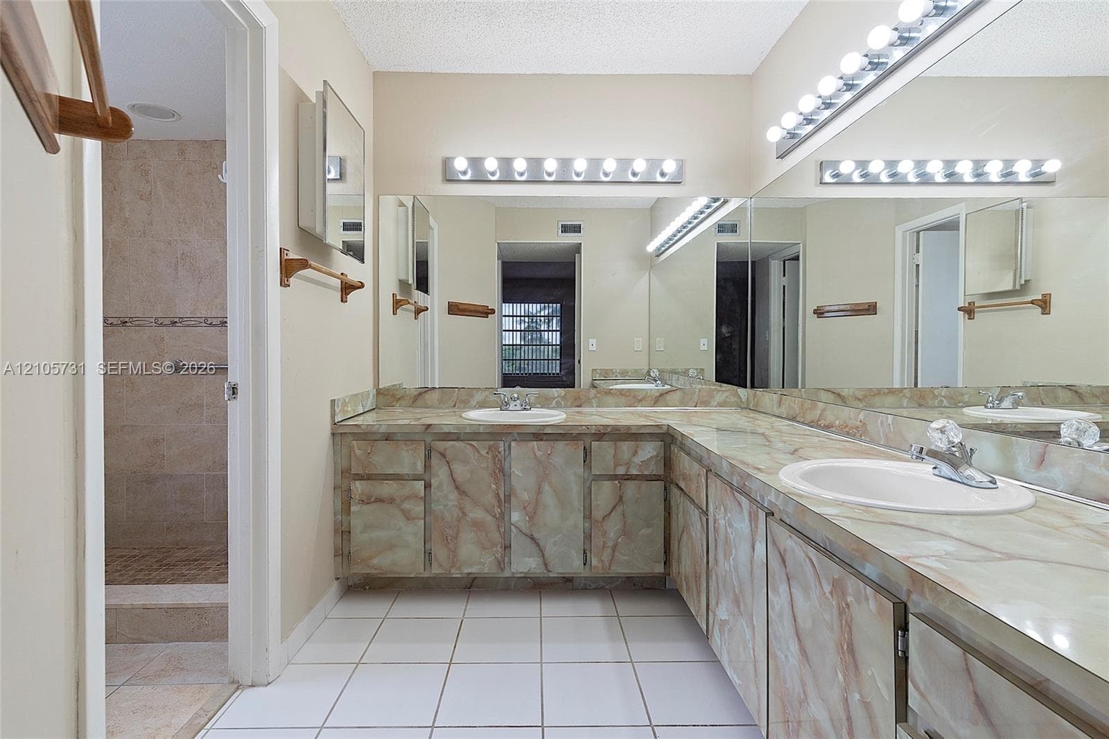 L-shaped vanity featuring custom-fitted stone-finish cabinetry and countertops, dual white drop-in sinks, extensive full-width wall mirrors, and integrated multi-bulb lighting fixtures