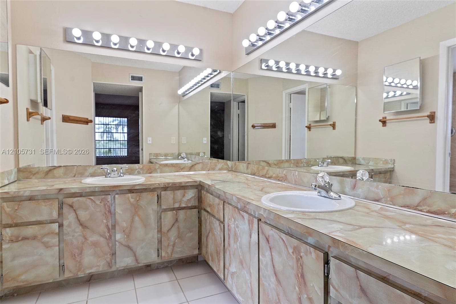 Dual-sink vanity featuring patterned stone-finish countertops, custom cabinetry with matching stone-finish doors, expansive wall-to-wall mirror, and multi-bulb theatrical lighting fixtures