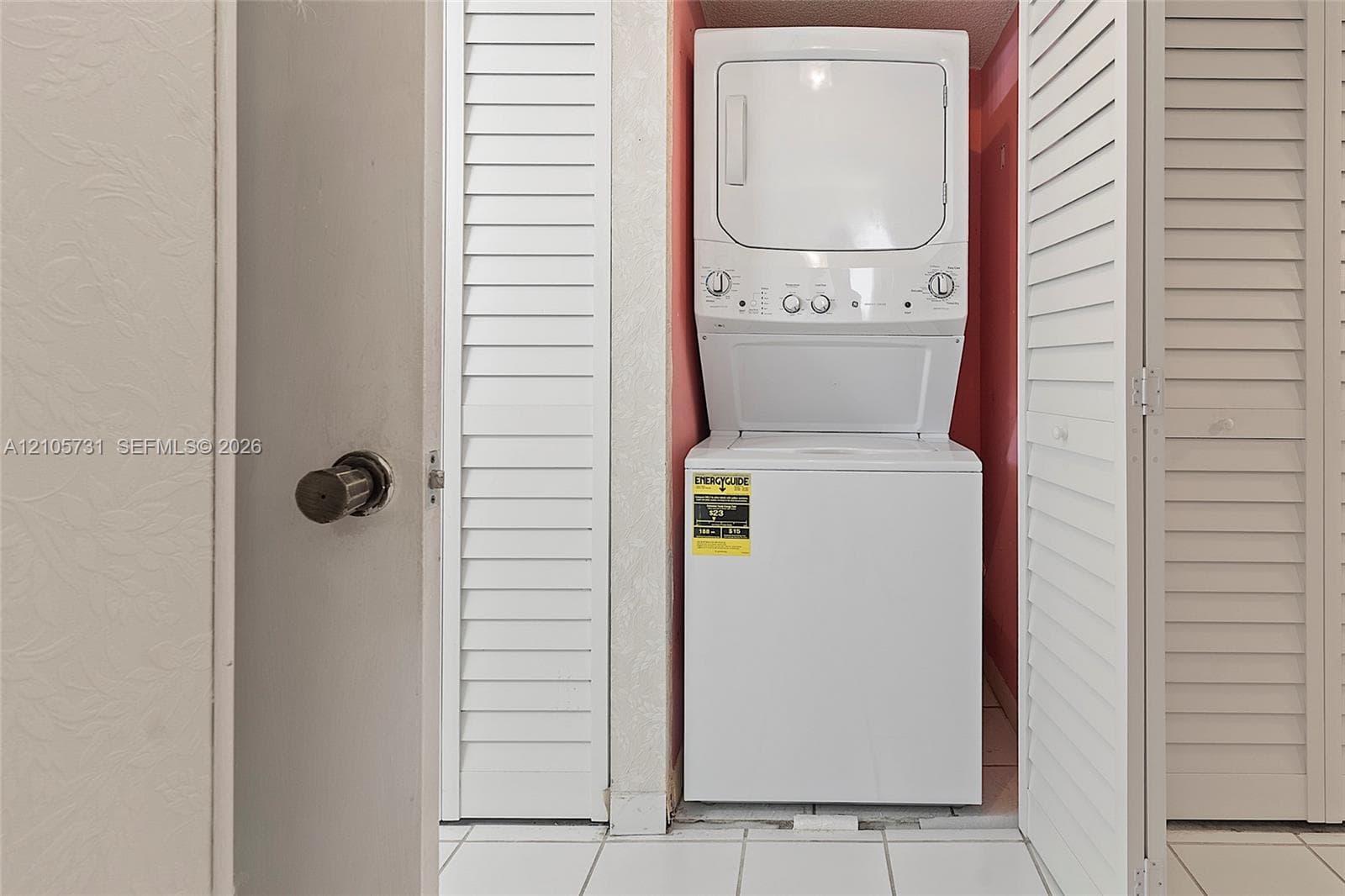 Stacked laundry unit housed within a closet featuring white louvered doors and tile flooring