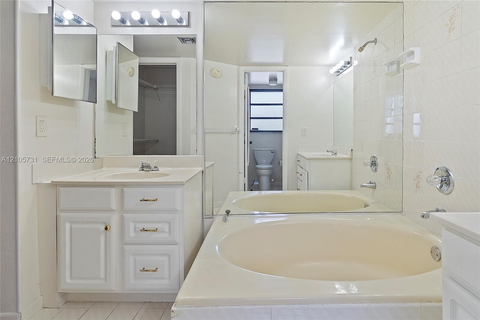 Bathroom featuring an oval soaking tub with a tiled surround, a white cabinetry vanity, an expansive wall-mounted mirror, and multi-bulb vanity lighting