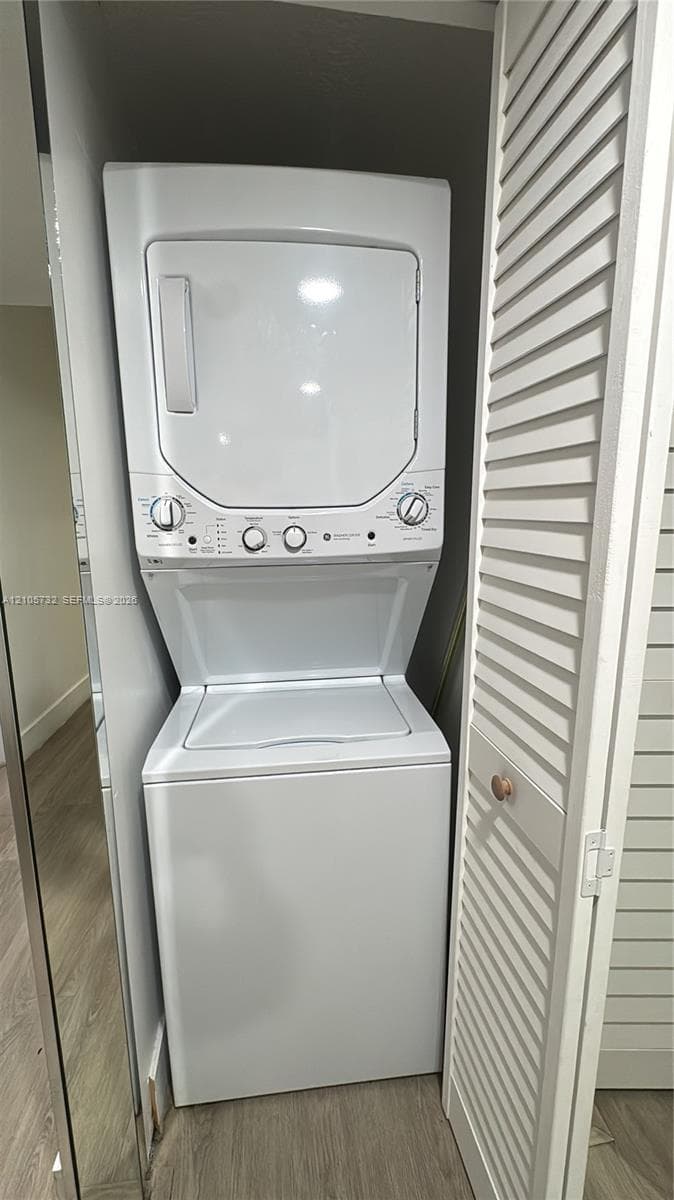 Stacked white washer and dryer unit housed within a closet featuring white louvered doors and wood-finish flooring