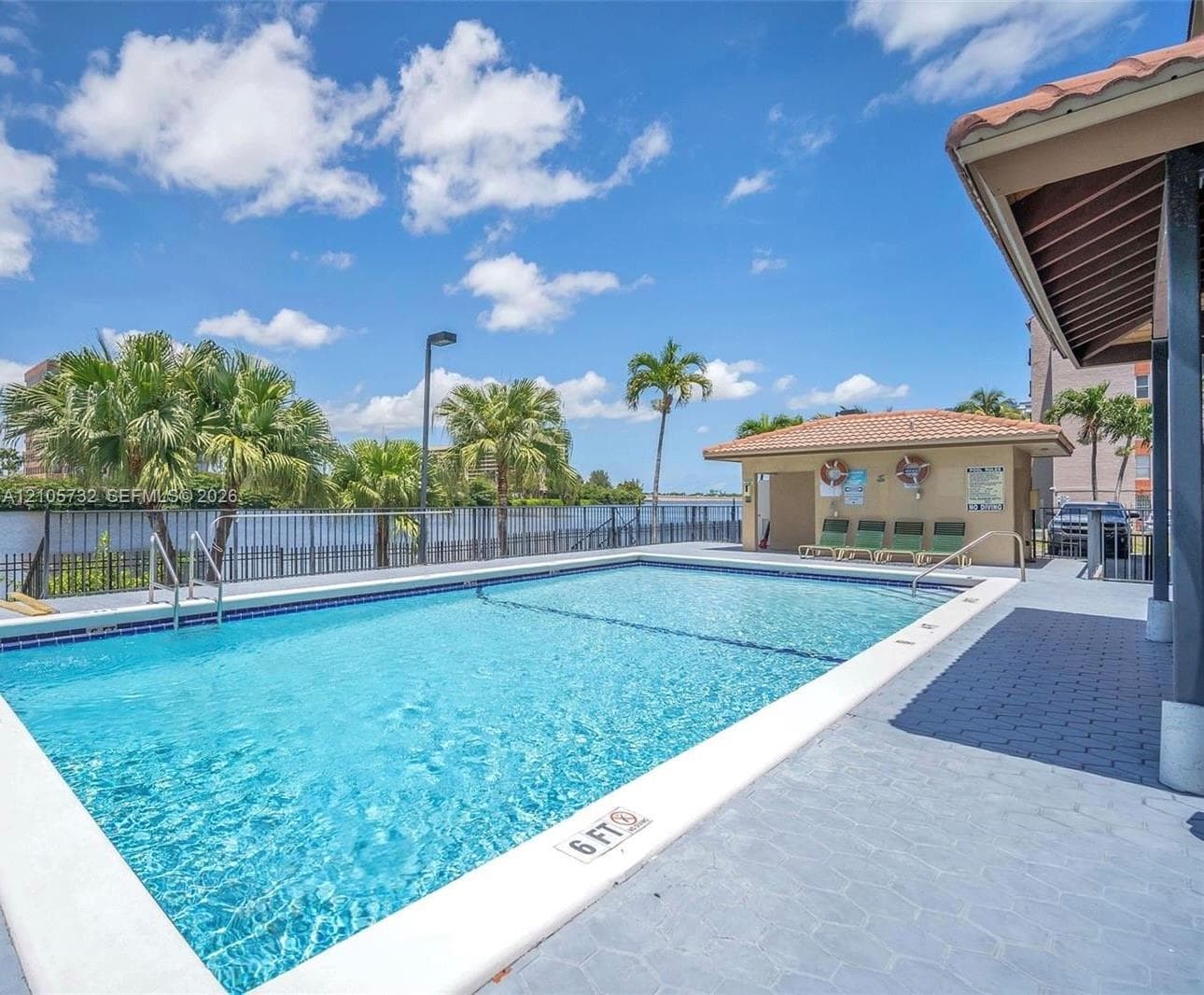 Outdoor swimming pool featuring a concrete deck, metal perimeter fencing, tiled roof cabana, and mature palm trees