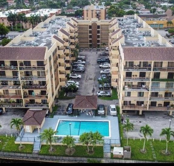 Aerial perspective of a multi-story residential complex featuring multiple private balconies, a ground-level swimming pool, a covered poolside pavilion, and paved surface parking