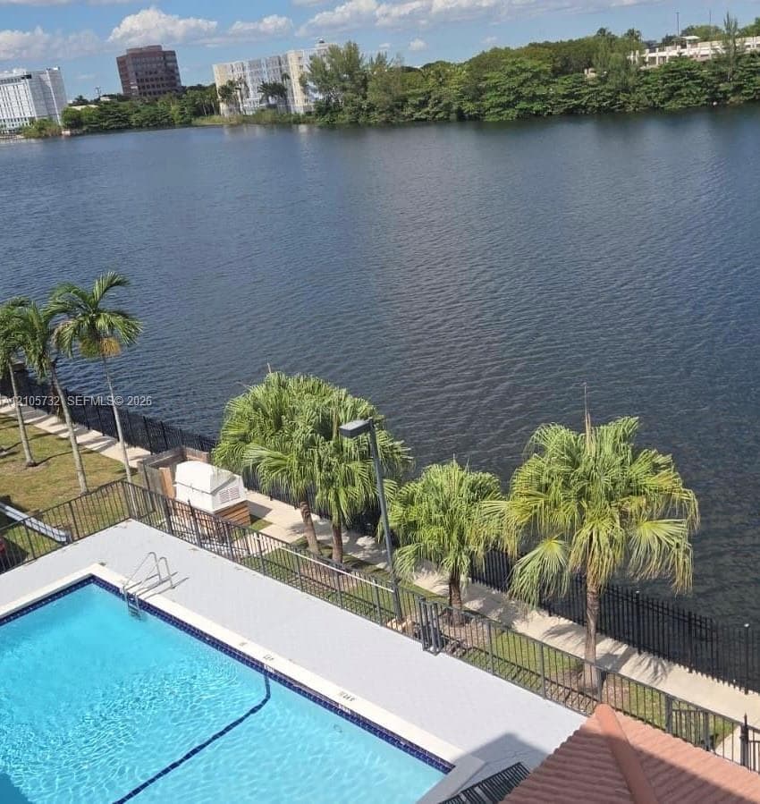 In-ground swimming pool with metal ladder, enclosed by black metal fencing, surrounded by palms and waterfront scenery