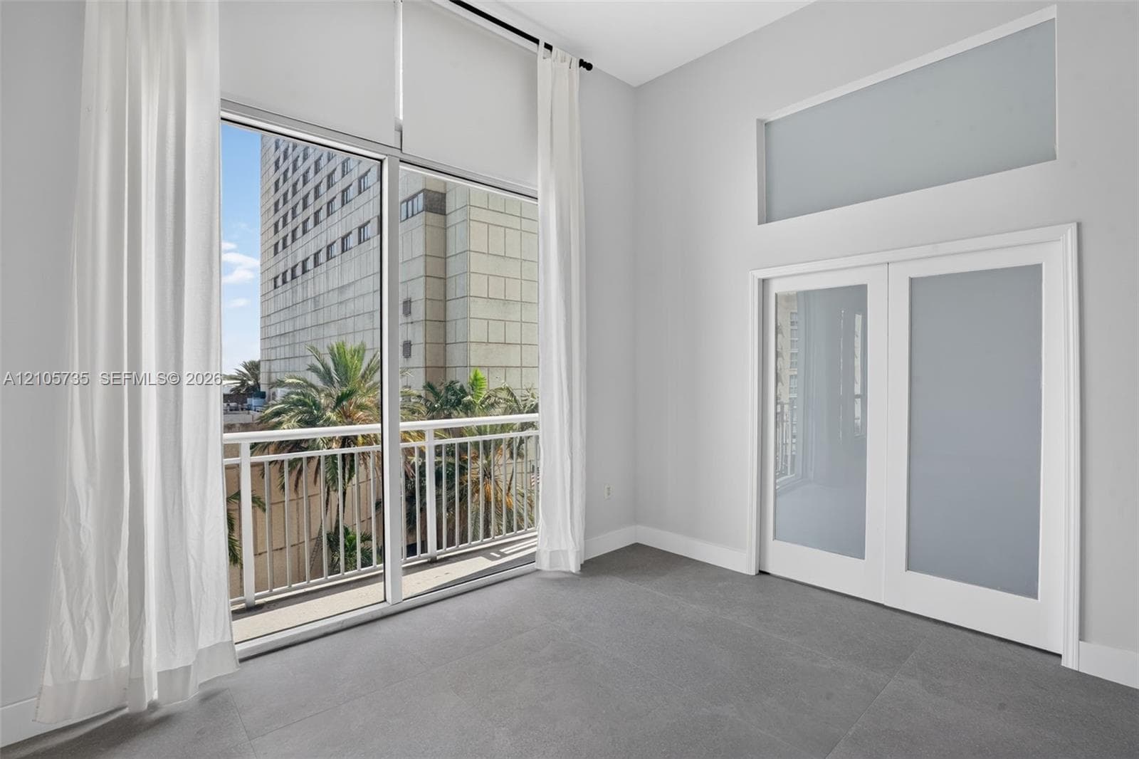 Floor-to-ceiling glass paneling, high ceilings, large-format tile flooring, a private balcony with metal railings, and dual French doors with frosted glass inserts