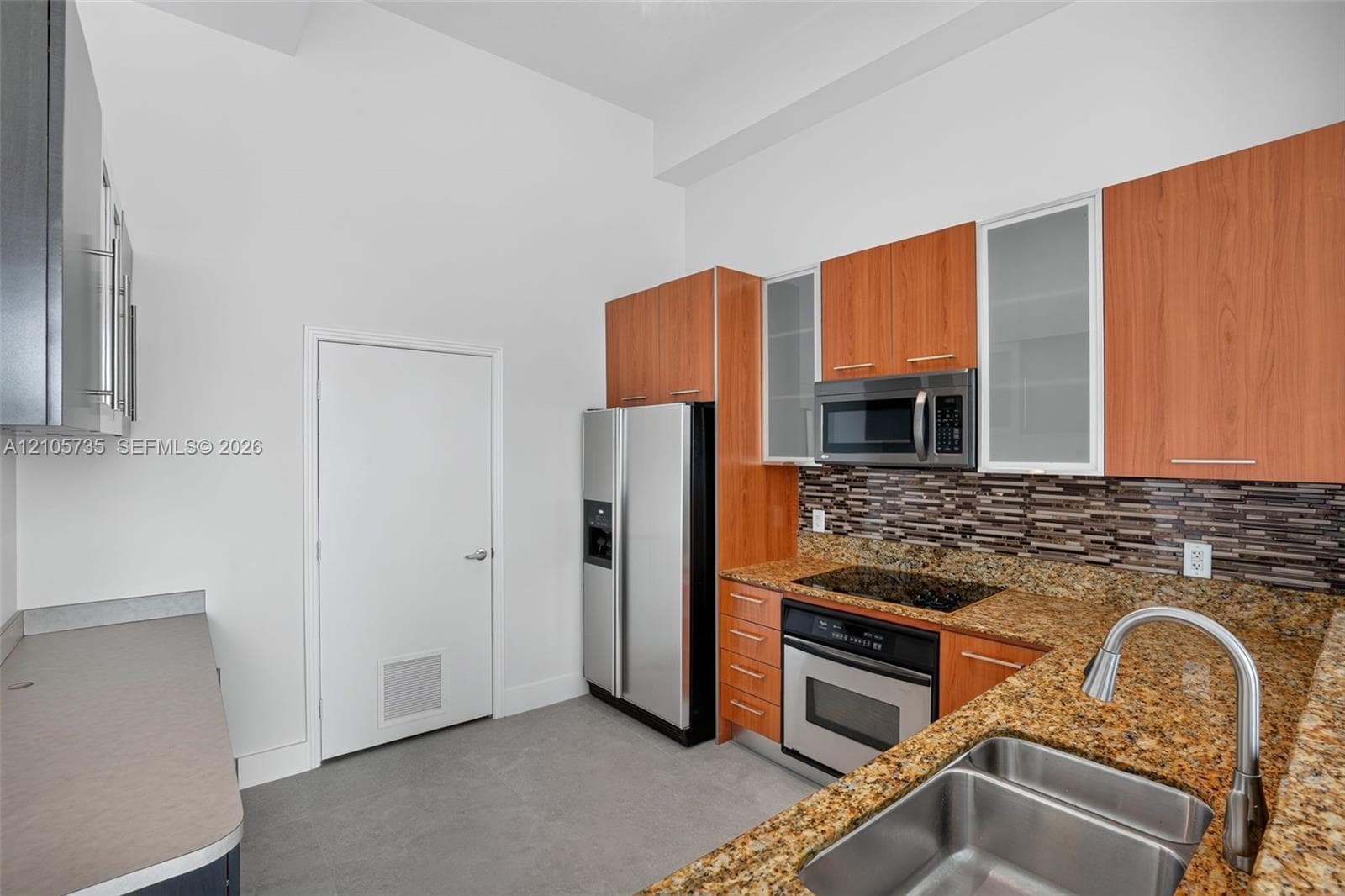 Kitchen featuring wood-finish cabinetry, stone-style countertops, stainless steel appliances, glass-front upper cabinet doors, and a multi-toned linear tile backsplash