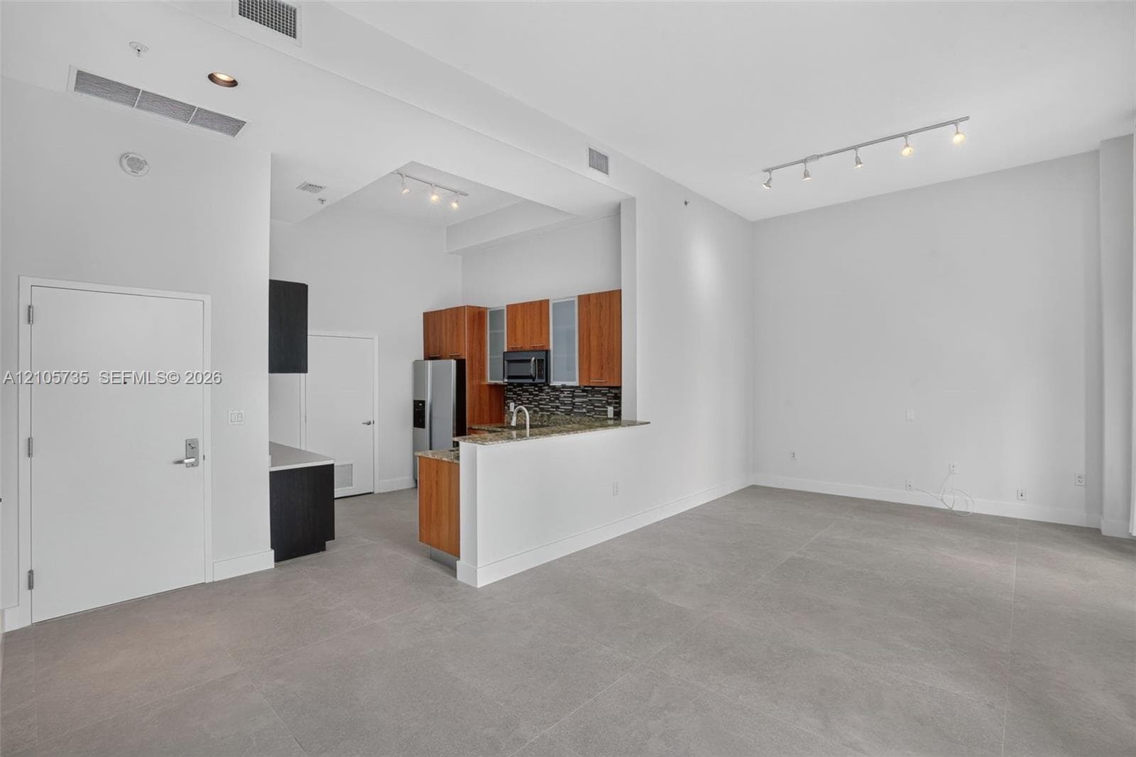 Open-concept interior featuring high ceilings, neutral tile flooring, track lighting, kitchen with wood-finish cabinetry, and stone-surface breakfast bar
