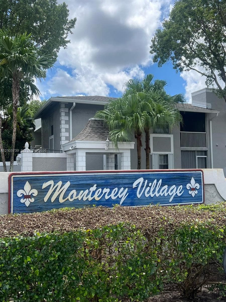 Community entrance featuring a monument sign, stucco-finished building facade, quoined corner detailing, and mature palm trees