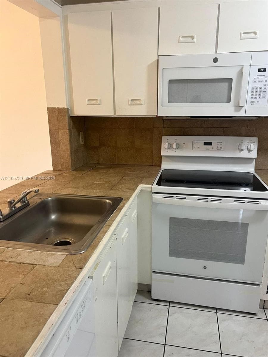 Kitchen featuring white cabinetry, tile countertops, tile backsplash, stainless steel sink, and white appliances including a built-in microwave and range