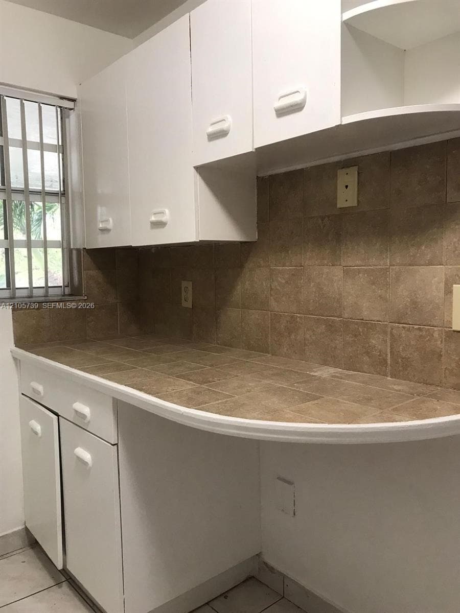 White cabinetry with built-in upper shelving, neutral tiled countertop, matching tile backsplash, and window with security bars