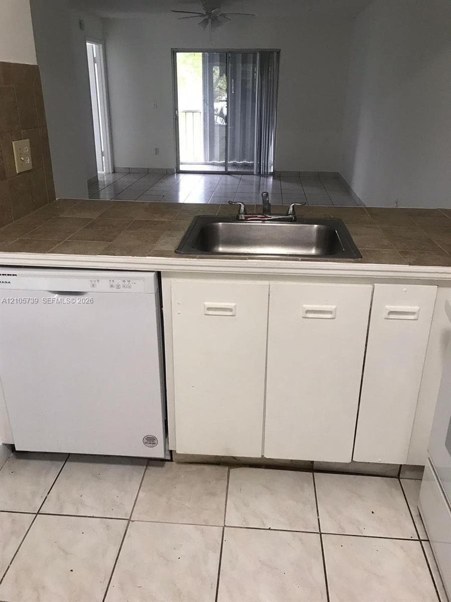 Kitchen featuring square tile countertops, undermount stainless steel sink with metal faucet, white dishwasher, white cabinetry, and ceramic tile flooring