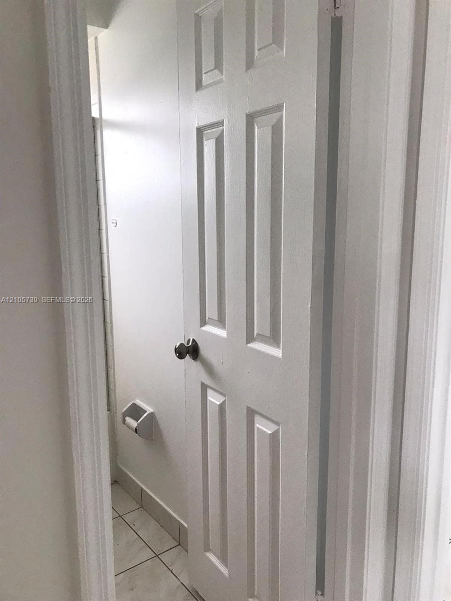 Six-panel door featuring a spherical brushed metal knob, wall-mounted toilet paper holder, and light-toned tile flooring