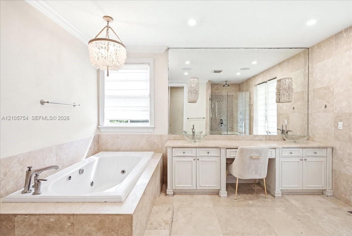 Jetted soaking tub with tile surround, double vanity with built-in dressing area, oversized wall mirror, decorative chandelier, and floor-to-ceiling tile wall finishes