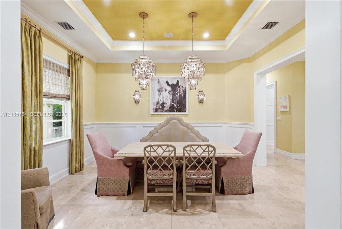 Tray ceiling featuring a decorative metallic finish, dual tiered crystal chandeliers, wall-mounted sconces, wainscoting, and stone-finish flooring