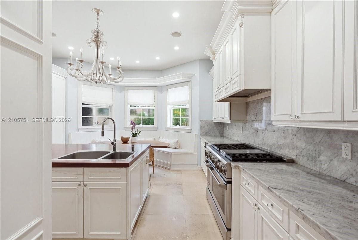 Kitchen featuring custom white cabinetry, stone countertops with matching backsplash, professional-grade stainless steel range, a large island with an integrated double-basin sink, and a built-in window seat