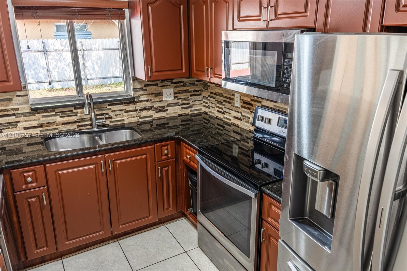 Kitchen featuring wood cabinetry, stone-finish countertops, stainless steel appliances, a multi-tone tile backsplash, and ceramic floor tiling