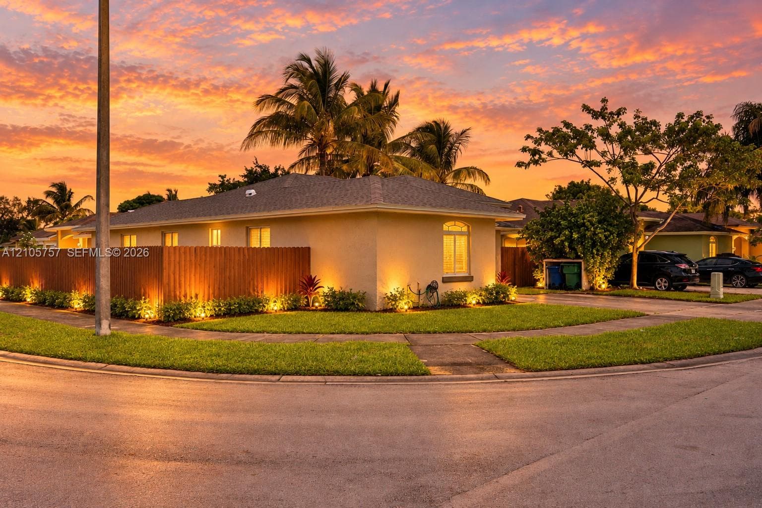 Single-story residence featuring a stucco exterior, integrated landscape lighting, a wood-panel perimeter fence, and established palm trees set against a prominent sunset backdrop