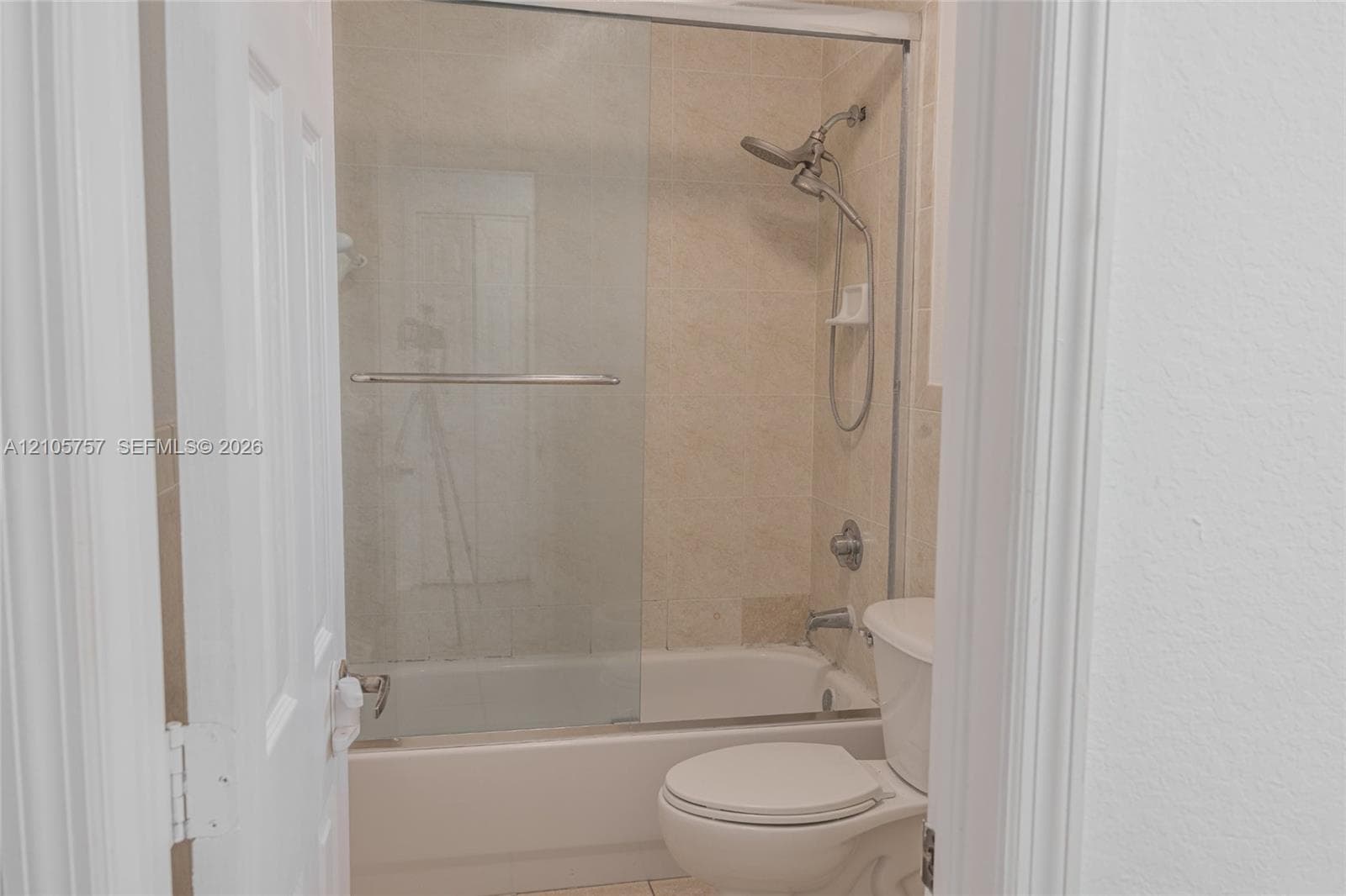 Bathroom featuring a tiled tub-shower combination, glass sliding enclosure, adjustable handheld shower fixture, and a white porcelain commode