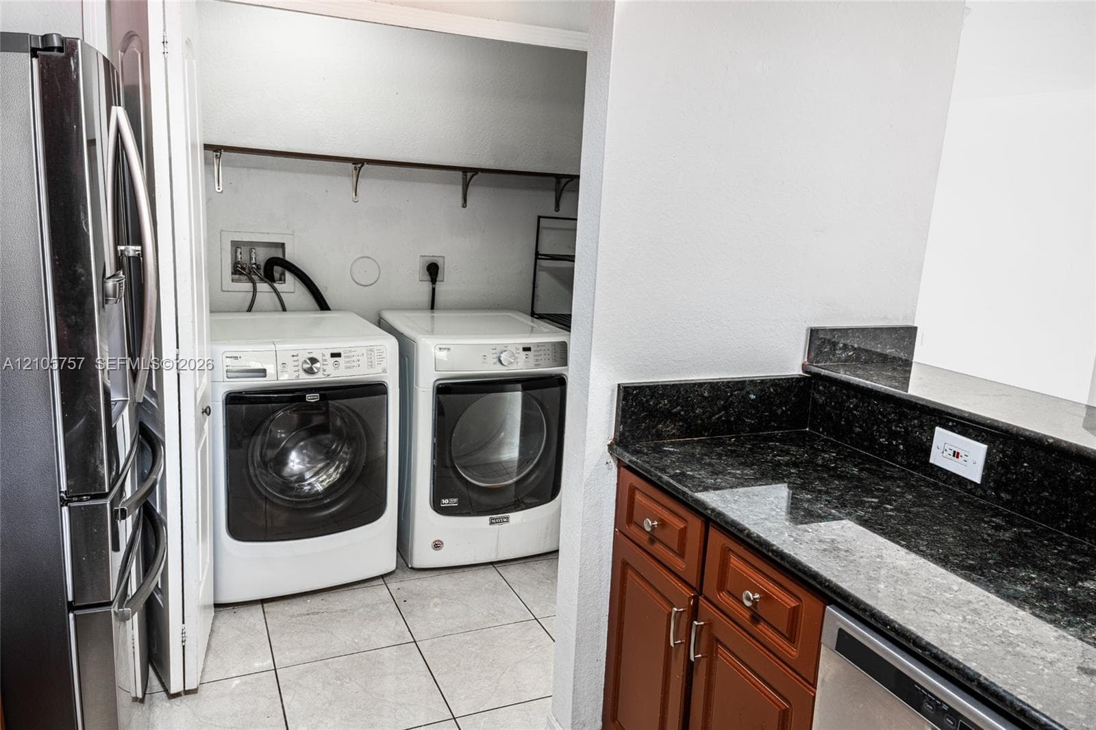 Laundry closet with built-in shelving, dark stone-finish countertops, warm-toned cabinetry, and tile flooring
