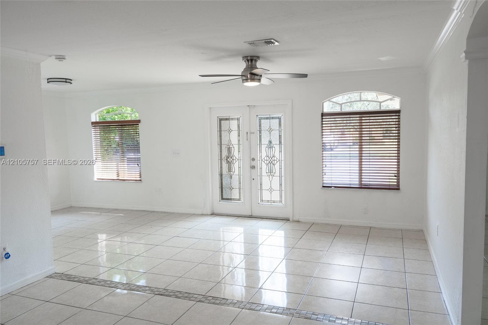 Living space featuring large-format tile flooring, dual French entry doors with decorative glass inserts, arched windows, crown molding, and a central ceiling fan