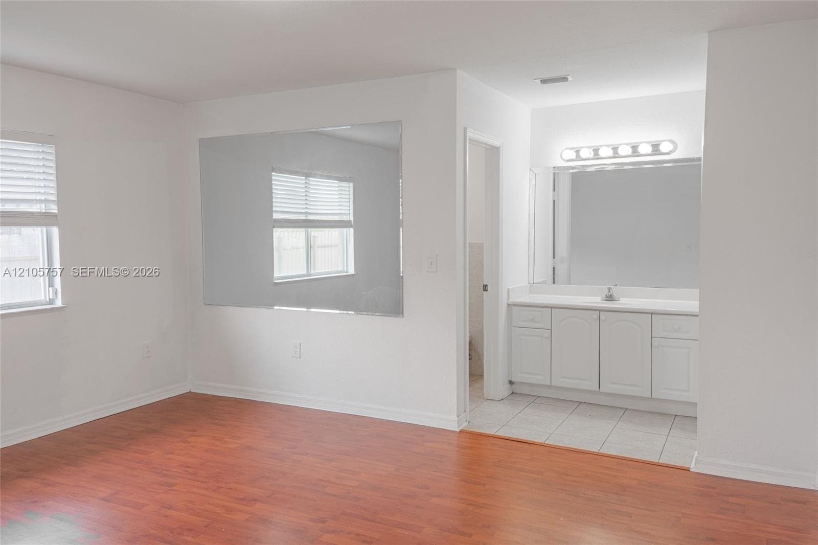 Wood-finish flooring, large wall-mounted mirror, white vanity cabinetry with surface-mount countertop, and horizontal slat window blinds