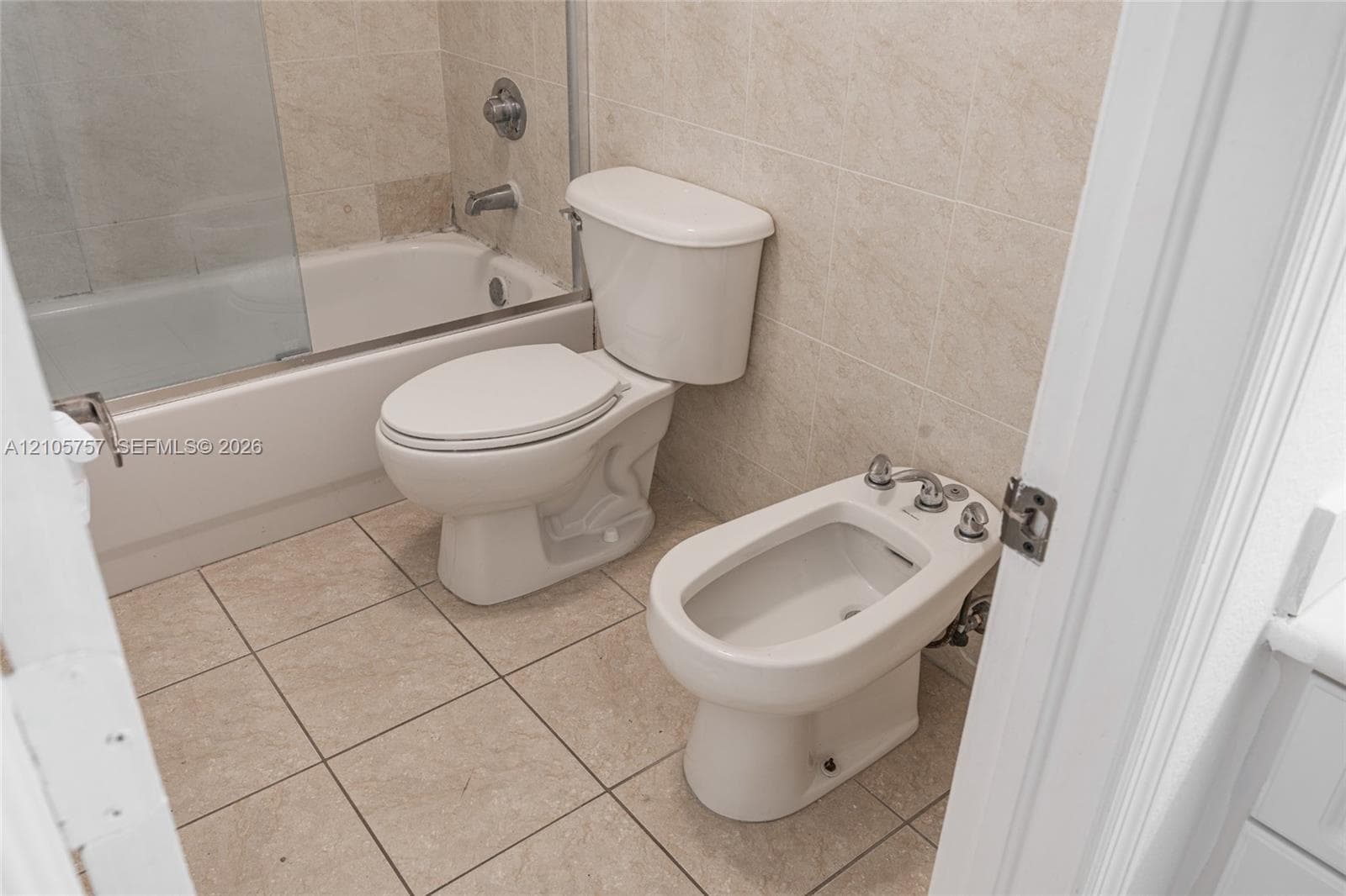 Full bathroom featuring a tub-shower combination with a glass door, tile wall surround, floor tiling, a standard toilet, and a bidet