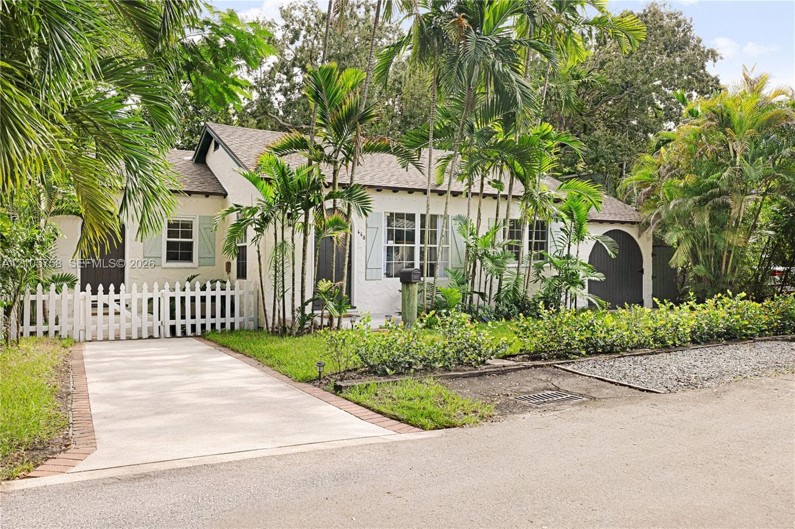 Stucco exterior featuring arched entry detailing, light-toned window shutters, a white picket fence, concrete driveway with brick edging, and mature palm landscaping