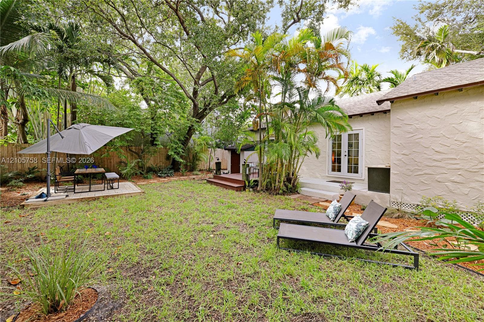 Rear exterior featuring a textured stucco facade, double glass-paneled doors, a raised wooden deck, a paved stone patio, and mature landscaping