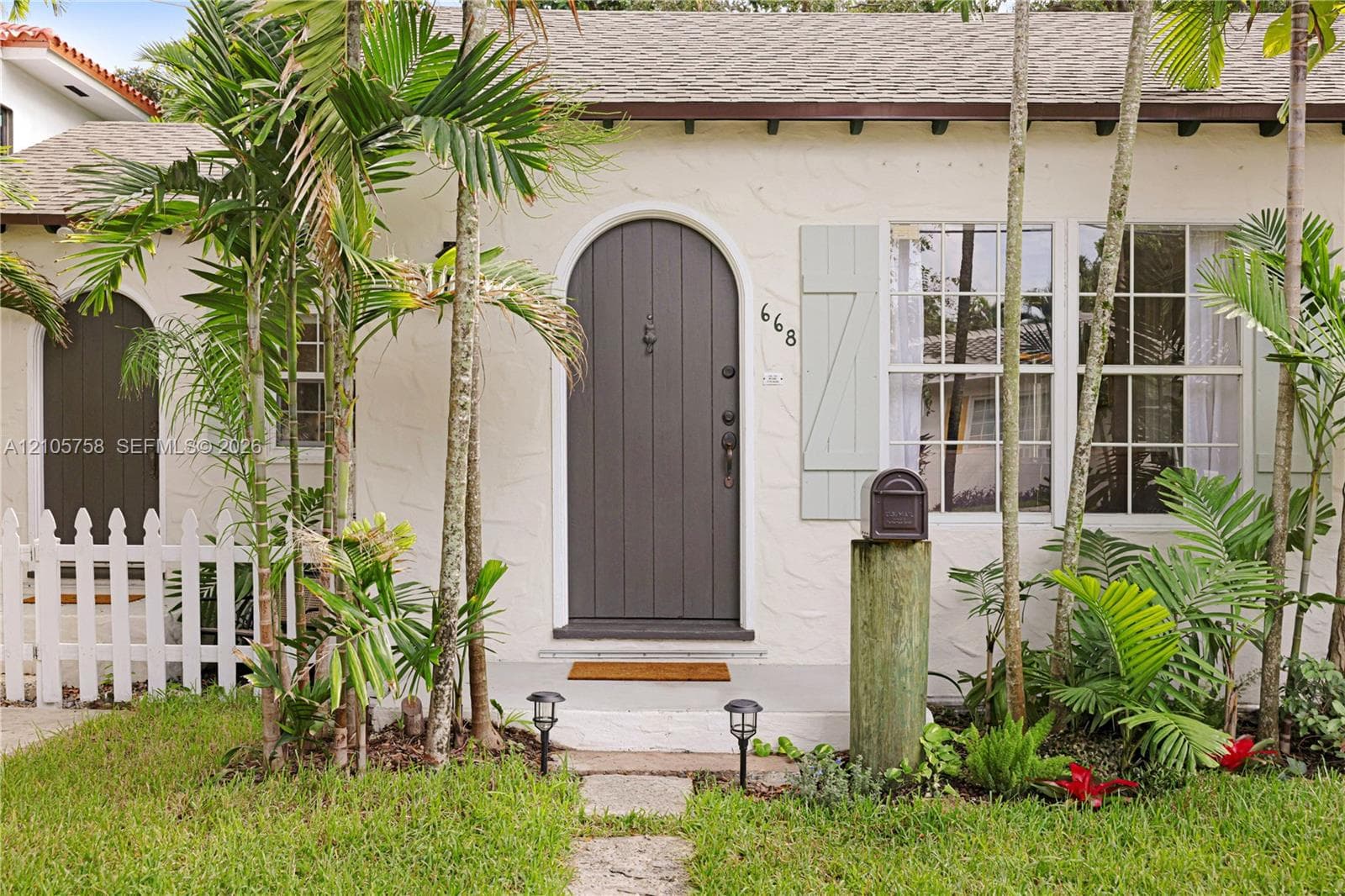 Exterior facade featuring stucco siding, an arched entry door, paneled shutters, and a white picket fence