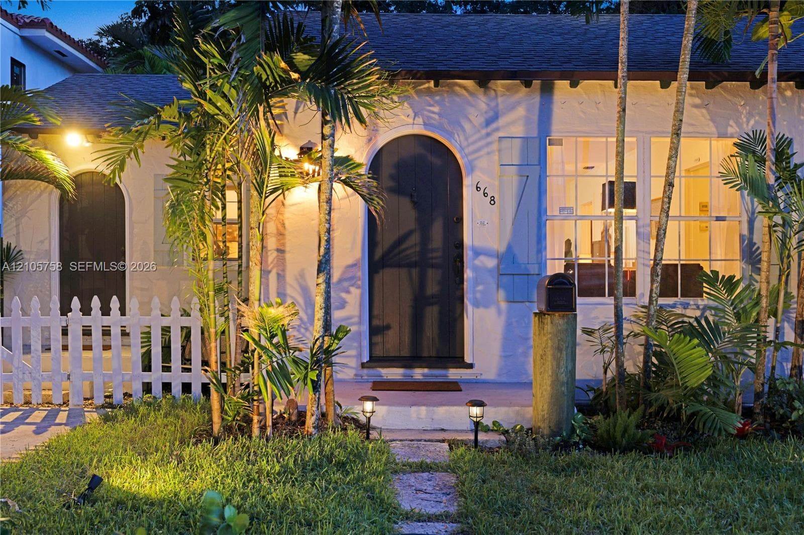 Stucco exterior featuring an arched entryway, shuttered multi-pane windows, a stone walkway, and a white picket fence