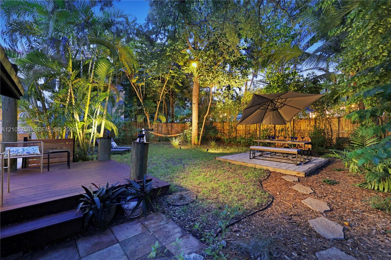 Backyard featuring a raised wood-finish deck, paved patio area, stone paver path, mature trees, and privacy fencing