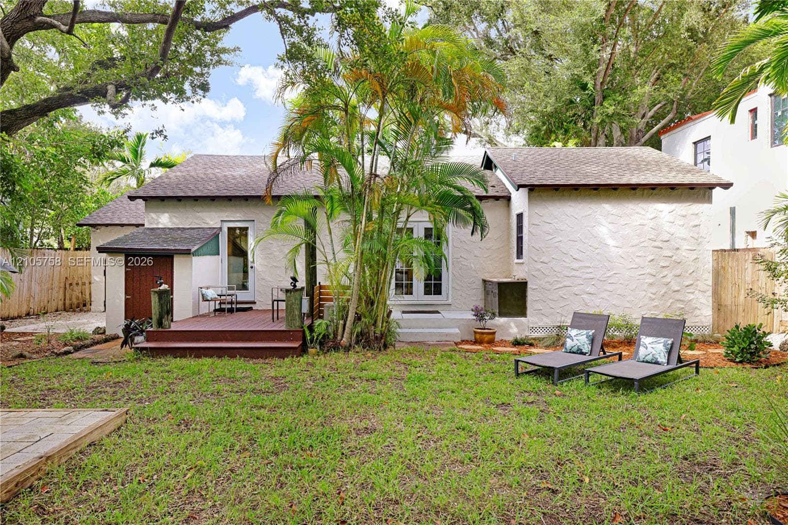Stucco exterior featuring a raised wood-finish deck, glass-paneled double doors, mature palm trees, and a fenced backyard
