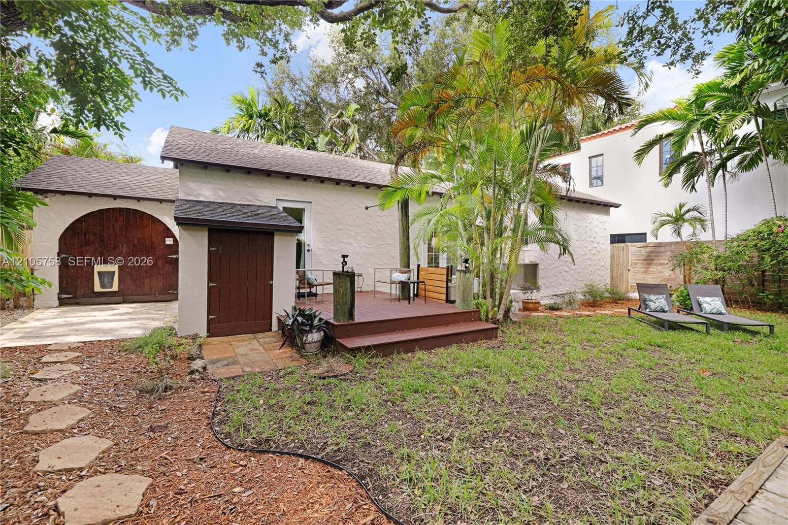 Stucco exterior featuring an arched wooden carriage door, raised wood-finish deck, stone paver pathway, and mature landscaping