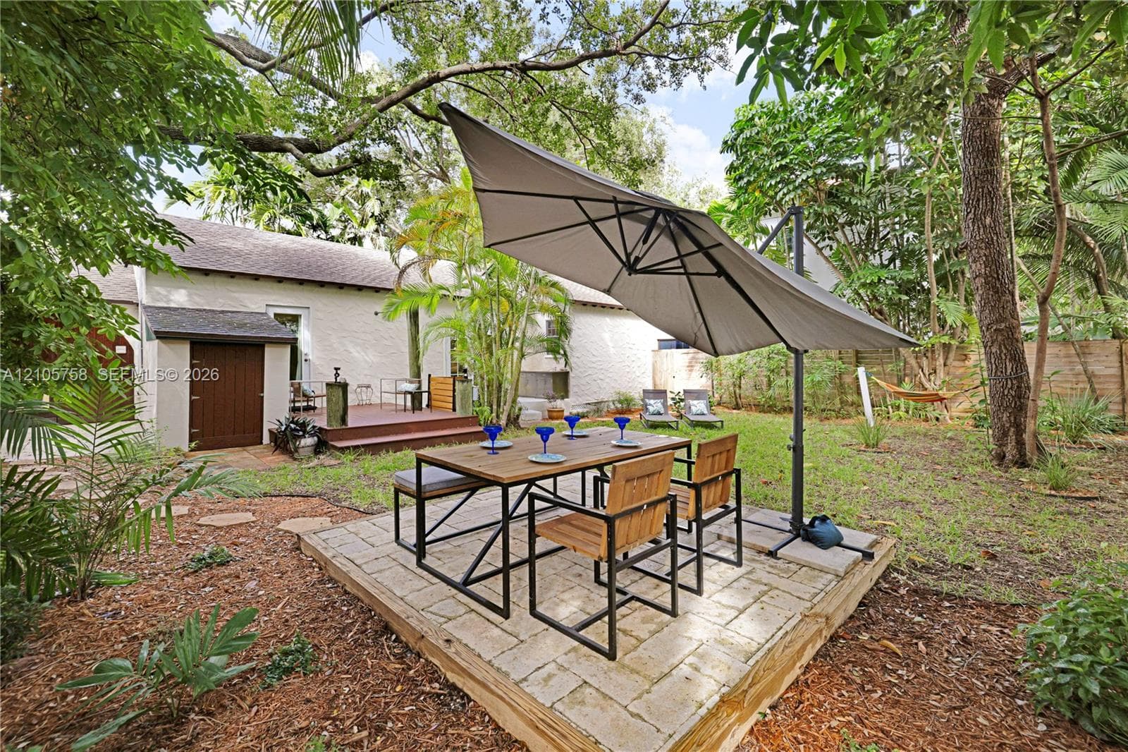 Backyard featuring a raised stone paver patio, elevated wood-decking platform, mature shade trees, and wood-finish exterior door