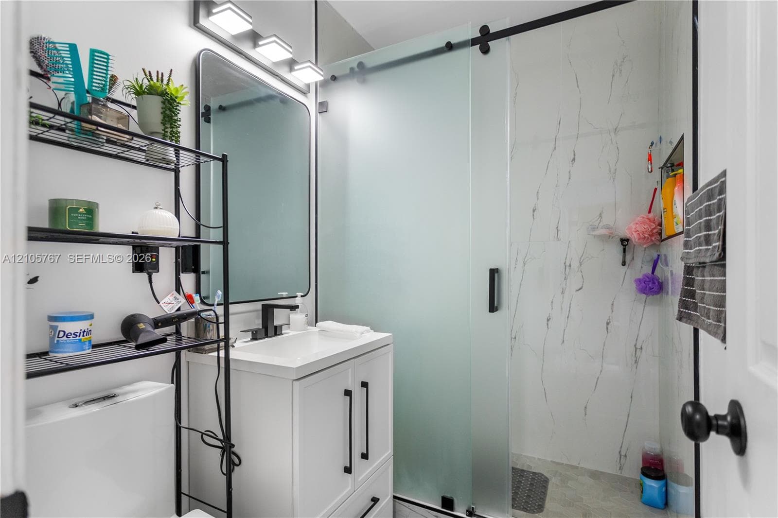 Bathroom featuring a white vanity cabinet with matte black hardware, a rectangular vanity sink, a frameless mirror with a matte black frame, and a walk-in shower with a sliding frosted glass door and wall tiles with a veined pattern