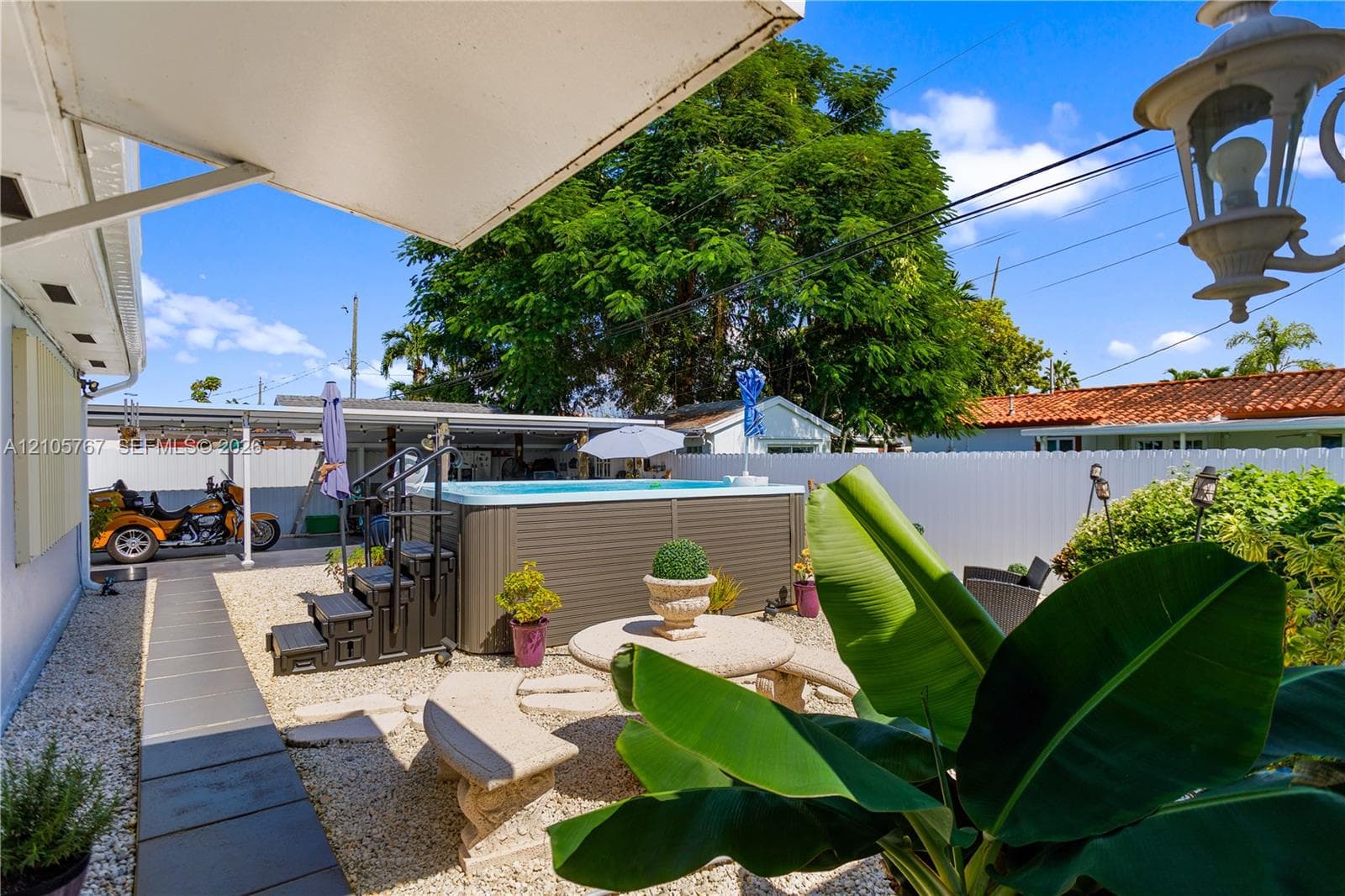 Rear exterior area featuring a freestanding hot tub, paved walkway, gravel ground cover, privacy fencing, and a built-in stone table with bench seating