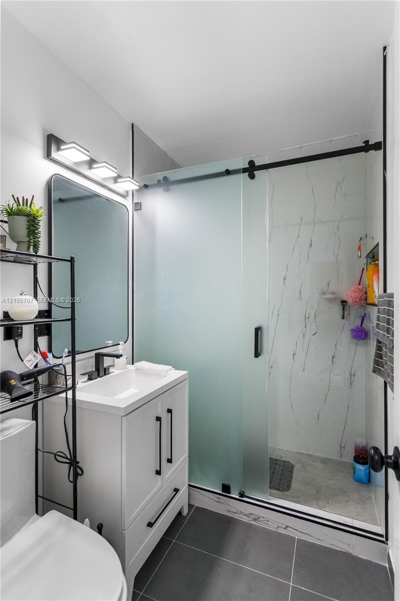 Modern bathroom featuring a white vanity with matte black hardware, a rectangular sink, a shower enclosure with frosted sliding glass doors and veined stone-style tile surround, and large-format gray floor tiles